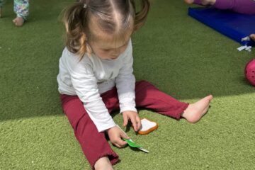 Petite fille assise sur une pelouse synthétique, jouant avec des accessoires de cuisine en plastique à la crèche Les Libellules