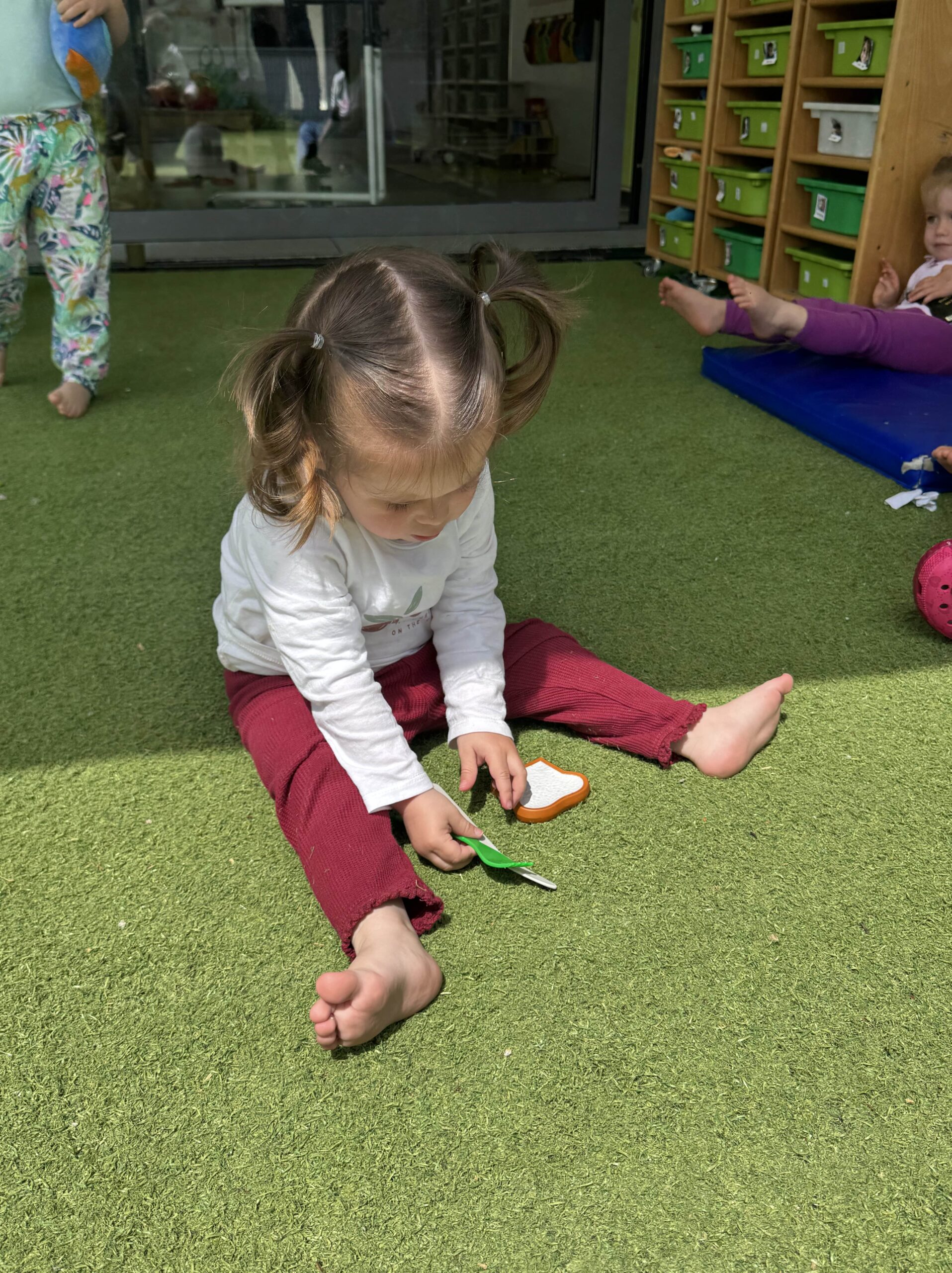 Petite fille assise sur une pelouse synthétique, jouant avec des accessoires de cuisine en plastique à la crèche Les Libellules