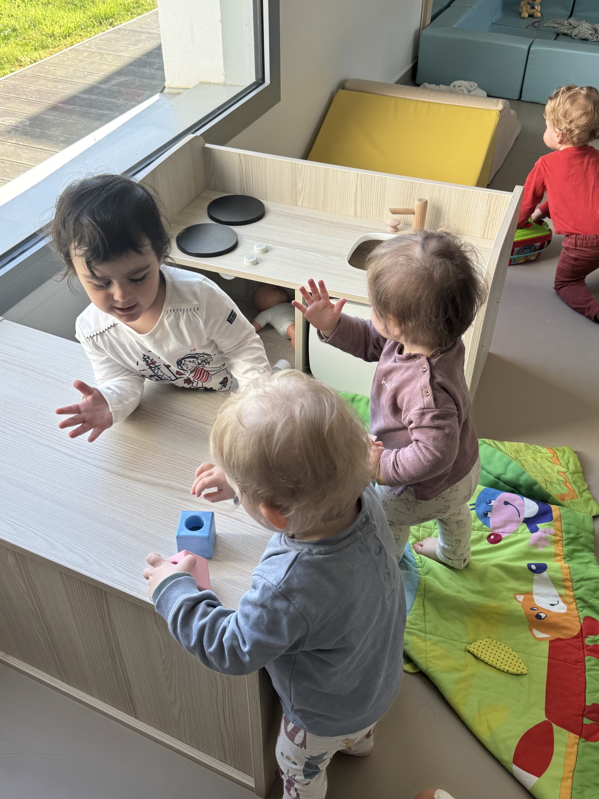 Trois jeunes enfants jouent ensemble autour d'une mini cuisine en bois dans la salle d'éveil à la crèche Les Papillons à Pontivy
