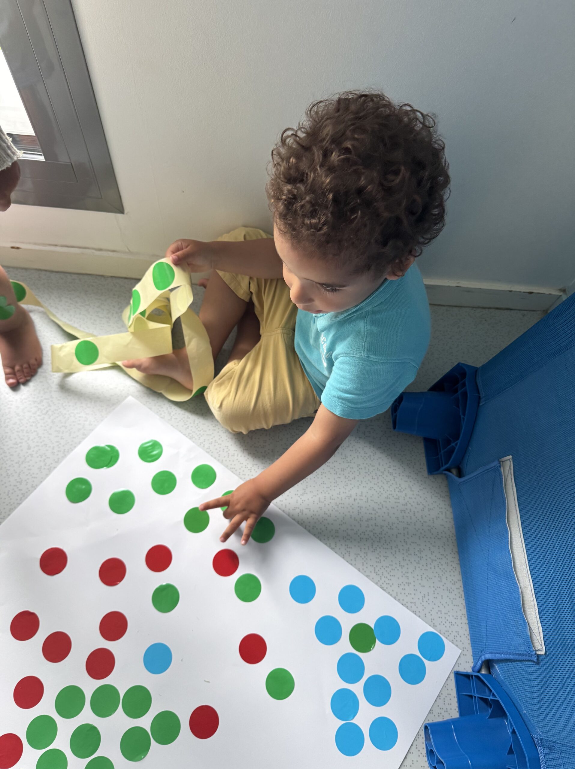 Petit garçon collant des gommettes colorés sur une grande feuille blanche à la crèche Les Libellules