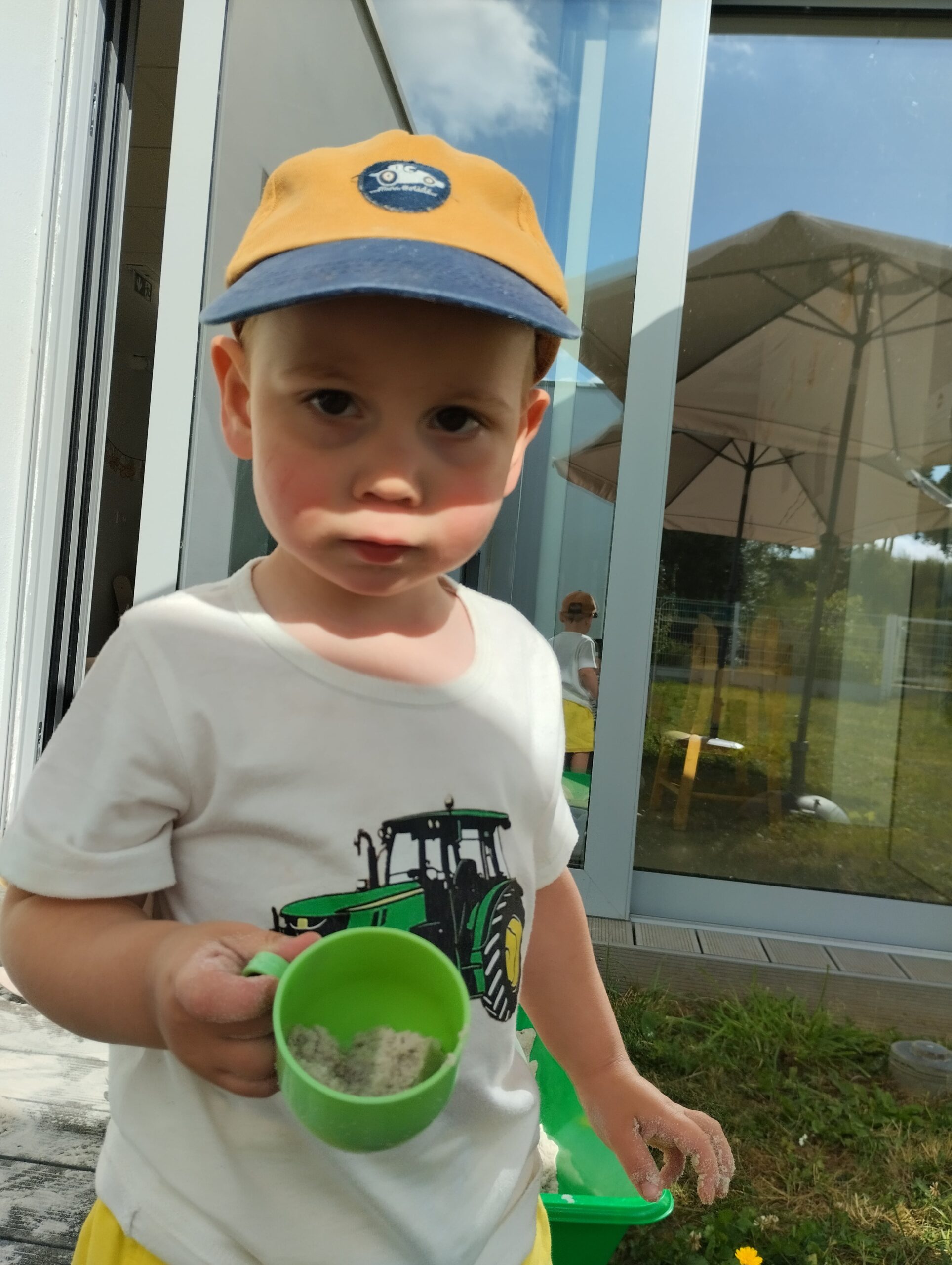 Petit garçon tenant une tasse verte remplie de sable devant la crèche Les Papillons à Pontivy.