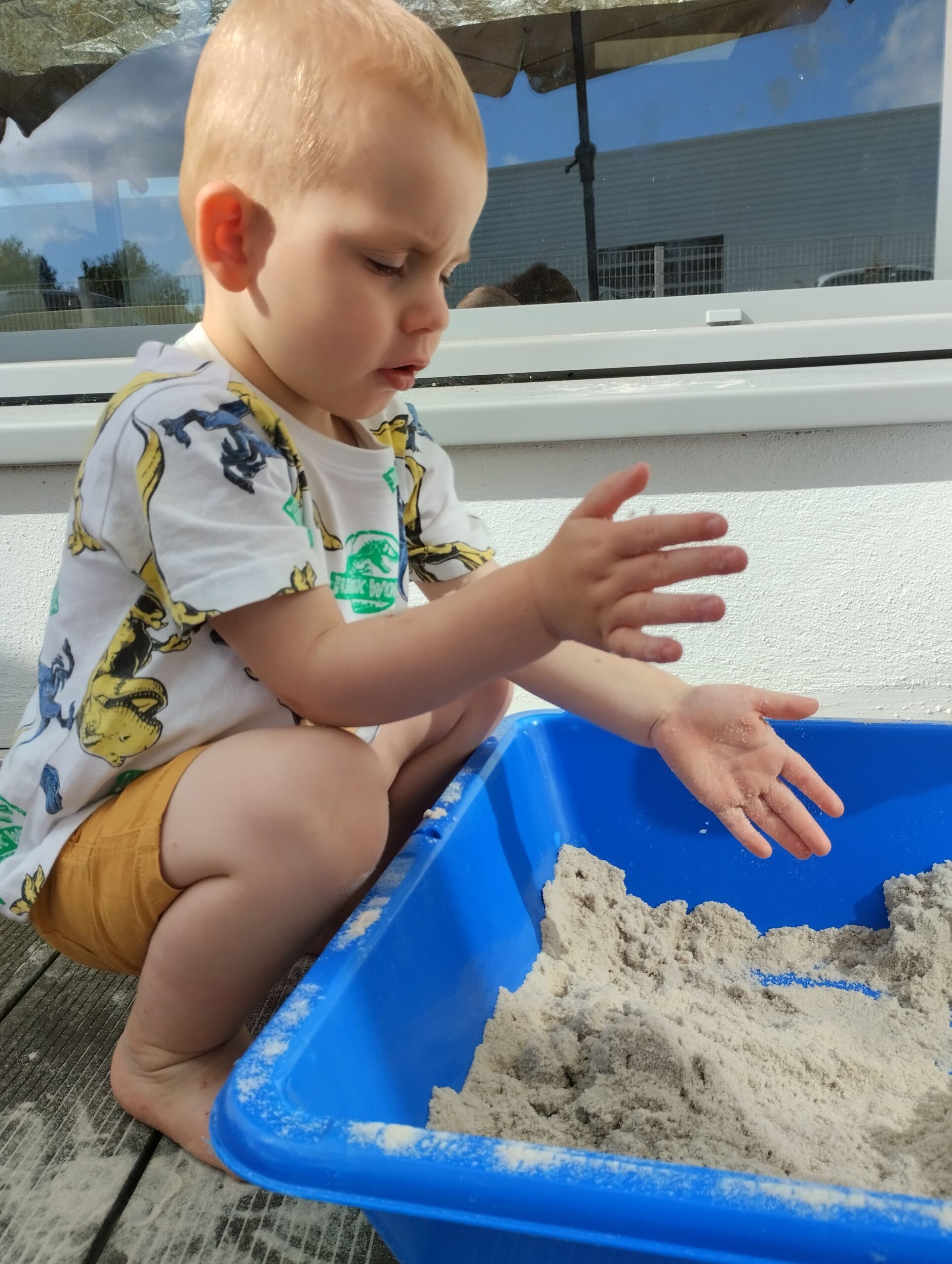 Tout-petit examinant ses mains pleines de sable, assis à côté d’un bac bleu à la crèche Les Papillons à Pontivy.
