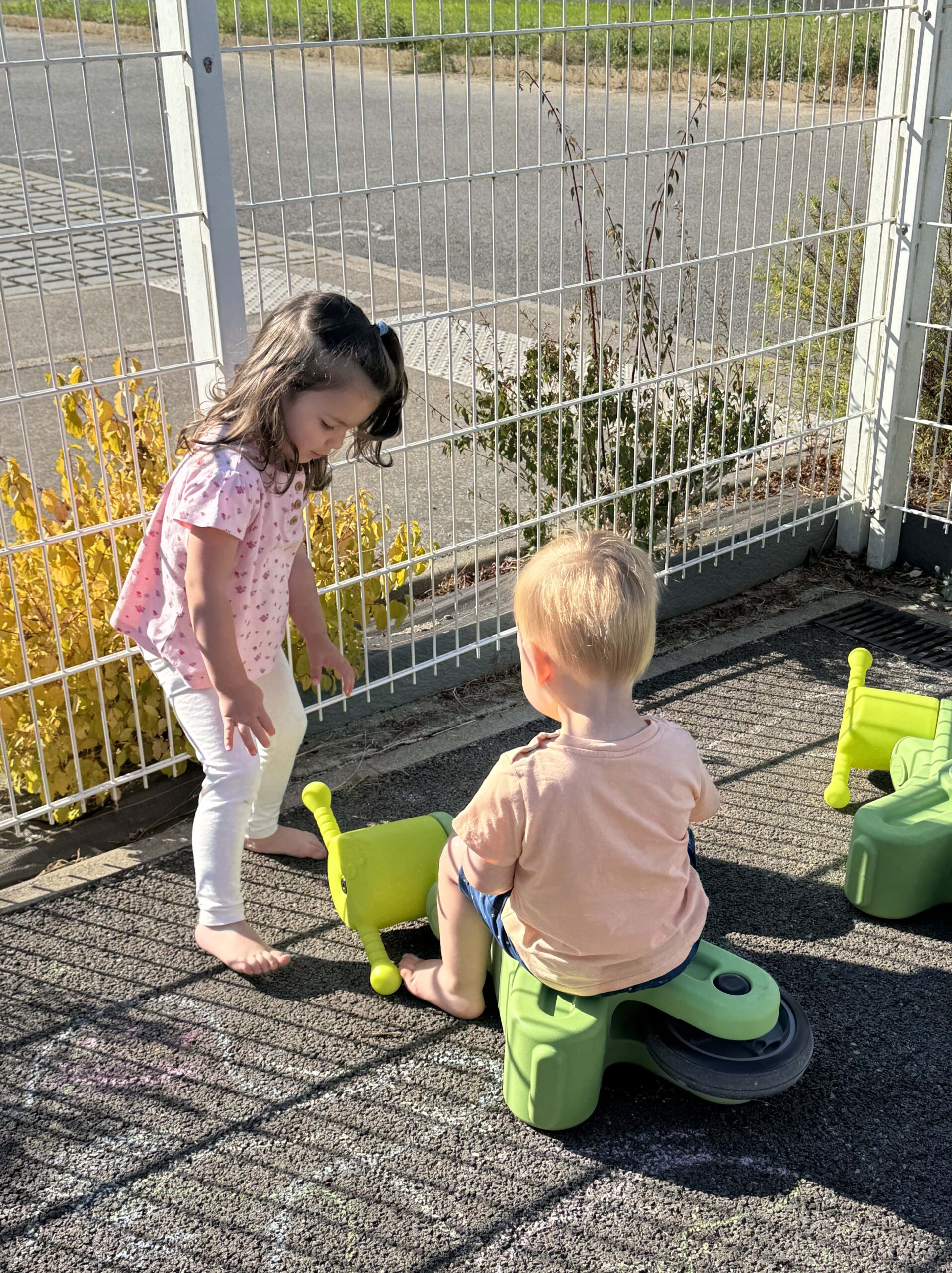 Deux enfants jouent ensemble sur un jouet vert, près de la clôture