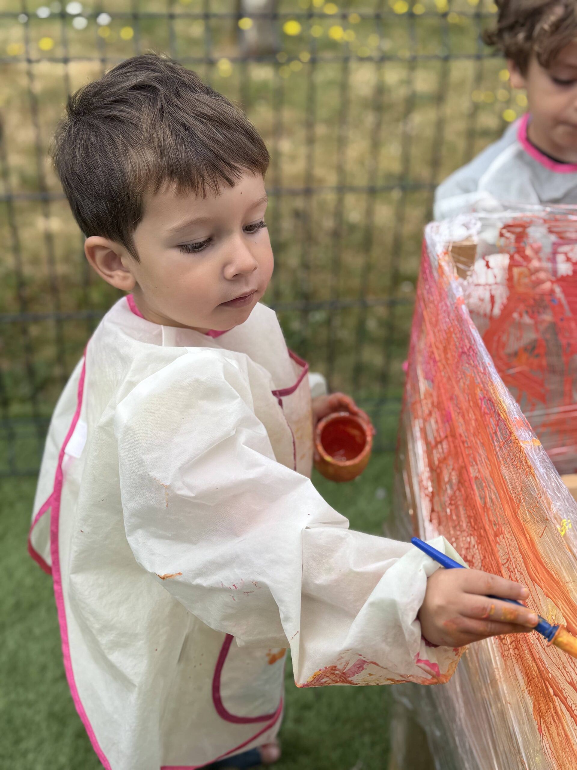 Enfant en blouse blanche concentré sur une activité de peinture en extérieur à la crèche Les Libellules