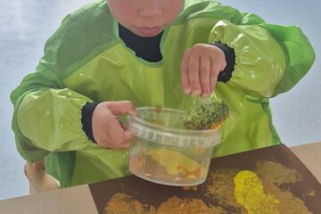 Un enfant en tablier vert réalise une activité de peinture à la crèche pour la Semaine du Goût en tamponnant de la peinture avec un morceau de brocoli sur une feuille de papier marron.