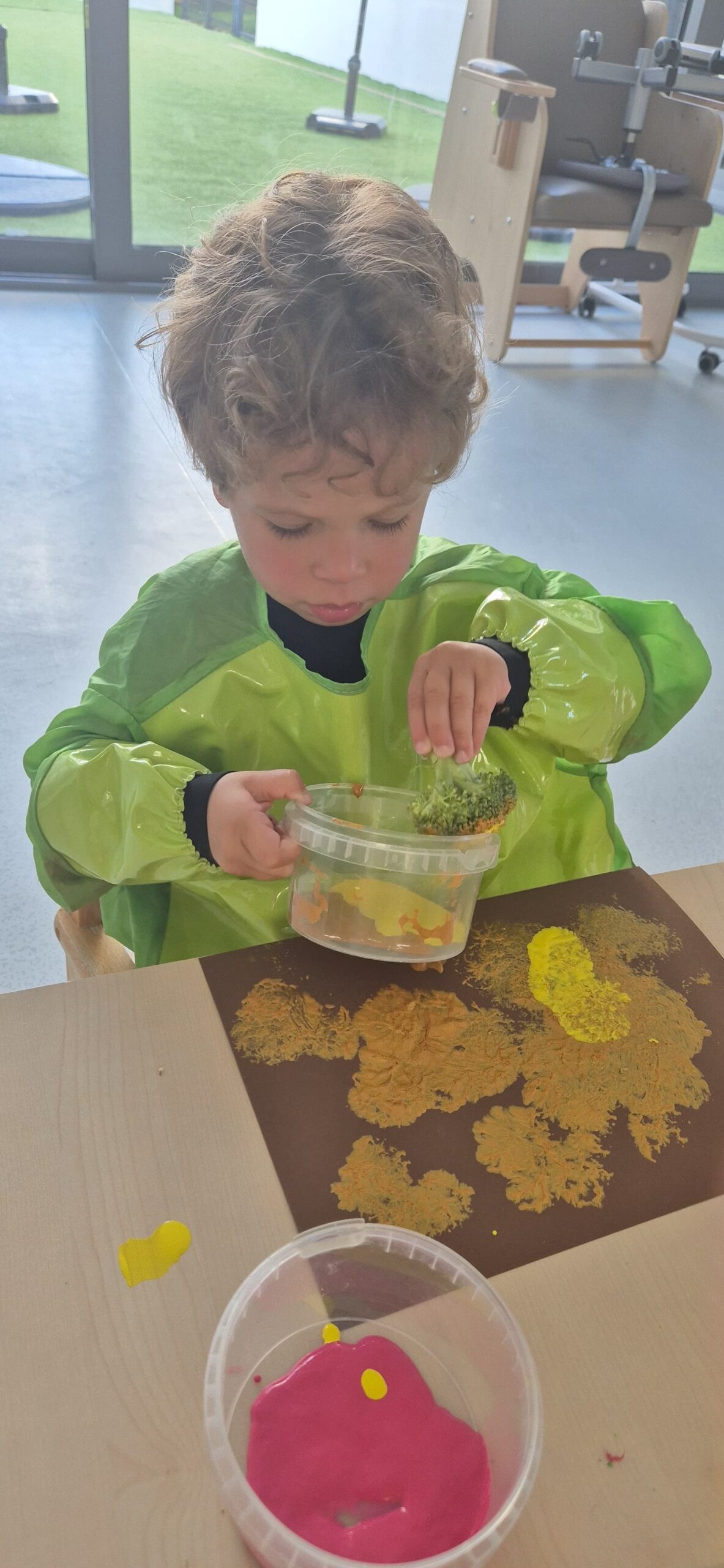 Un enfant en tablier vert réalise une activité de peinture à la crèche pour la Semaine du Goût en tamponnant de la peinture avec un morceau de brocoli sur une feuille de papier marron.