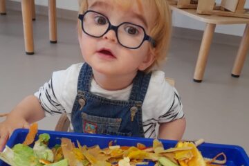 Un jeune enfant portant des lunettes et une salopette observe un plateau rempli d'épluchures et de morceaux de fruits et légumes colorés lors d'un atelier sensoriel à la crèche pendant la Semaine du goût.
