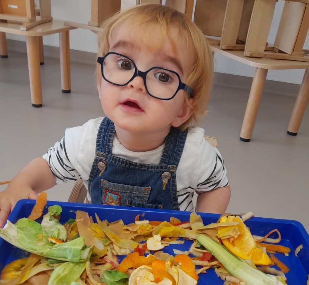Un jeune enfant portant des lunettes et une salopette observe un plateau rempli d'épluchures et de morceaux de fruits et légumes colorés lors d'un atelier sensoriel à la crèche pendant la Semaine du goût.