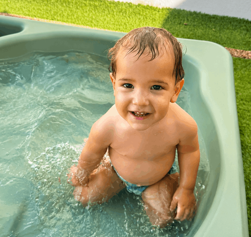 Un jeune enfant souriant est assis dans une petite piscine en plastique vert de la crèche Les Libellules, en train de jouer dans l'eau. Le bassin est posé sur de la pelouse artificielle.