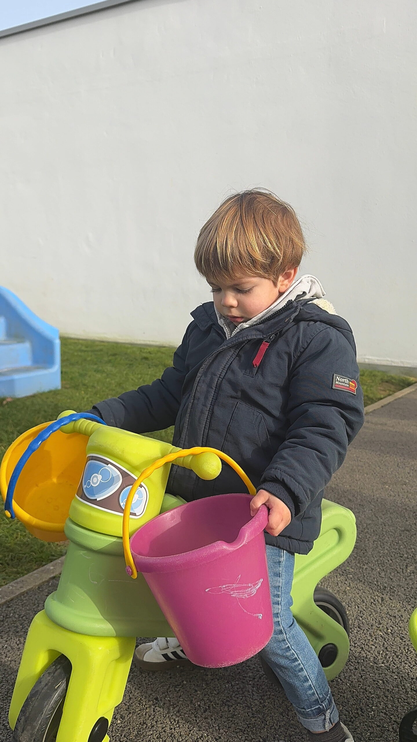 À la crèche Les Papillons à Pontivy, un enfant en manteau bleu marine est assis sur un tricycle vert fluo. Il tient fermement le guidon auquel sont suspendus deux seaux en plastique : un jaune et un violet. Il regarde sur le côté. L'arrière-plan montre la pelouse et le bâtiment de la crèche Les Papillons.