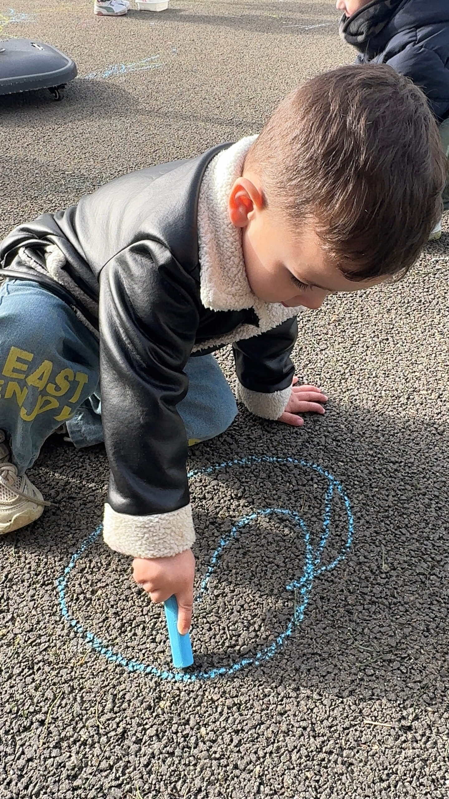 Dans la cour de la crèche Les Papillons à Pontivy, un enfant portant une veste en cuir à col en peau lainée est agenouillé sur le bitume. Il est concentré, tenant une grosse craie bleue avec laquelle il trace une spirale sur le sol. L'arrière-plan montre les autres enfants et les jeux de la crèche Les Papillons.