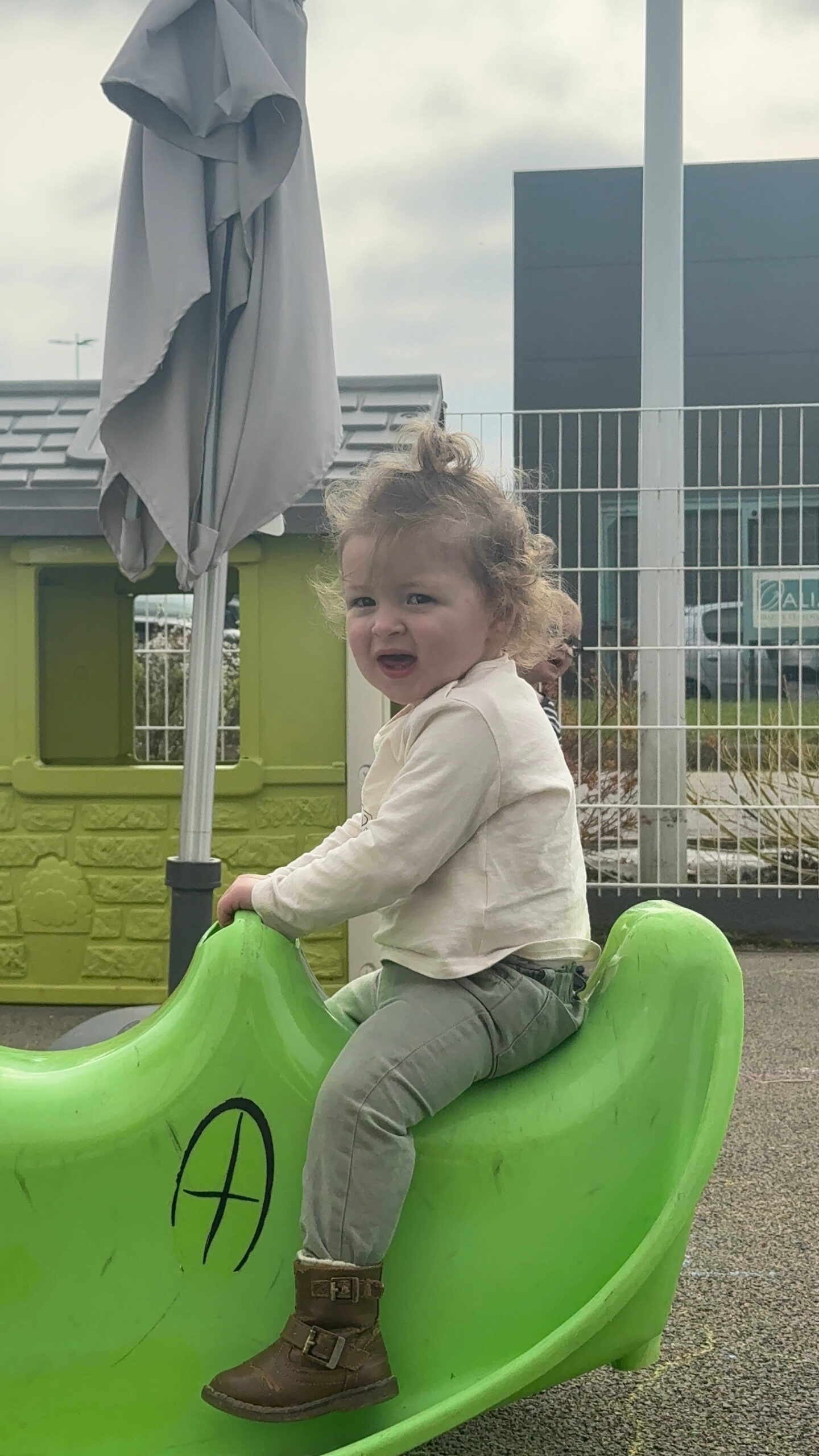 Dans le jardin de la crèche Les Papillons à Pontivy, une petite fille aux cheveux bouclés, portant un sweat-shirt beige et un pantalon vert, est assise sur un jeu à bascule en plastique vert. Elle sourit et regarde l'objectif. Derrière elle, on aperçoit une autre enfant et les installations de la crèche Les Papillons.
