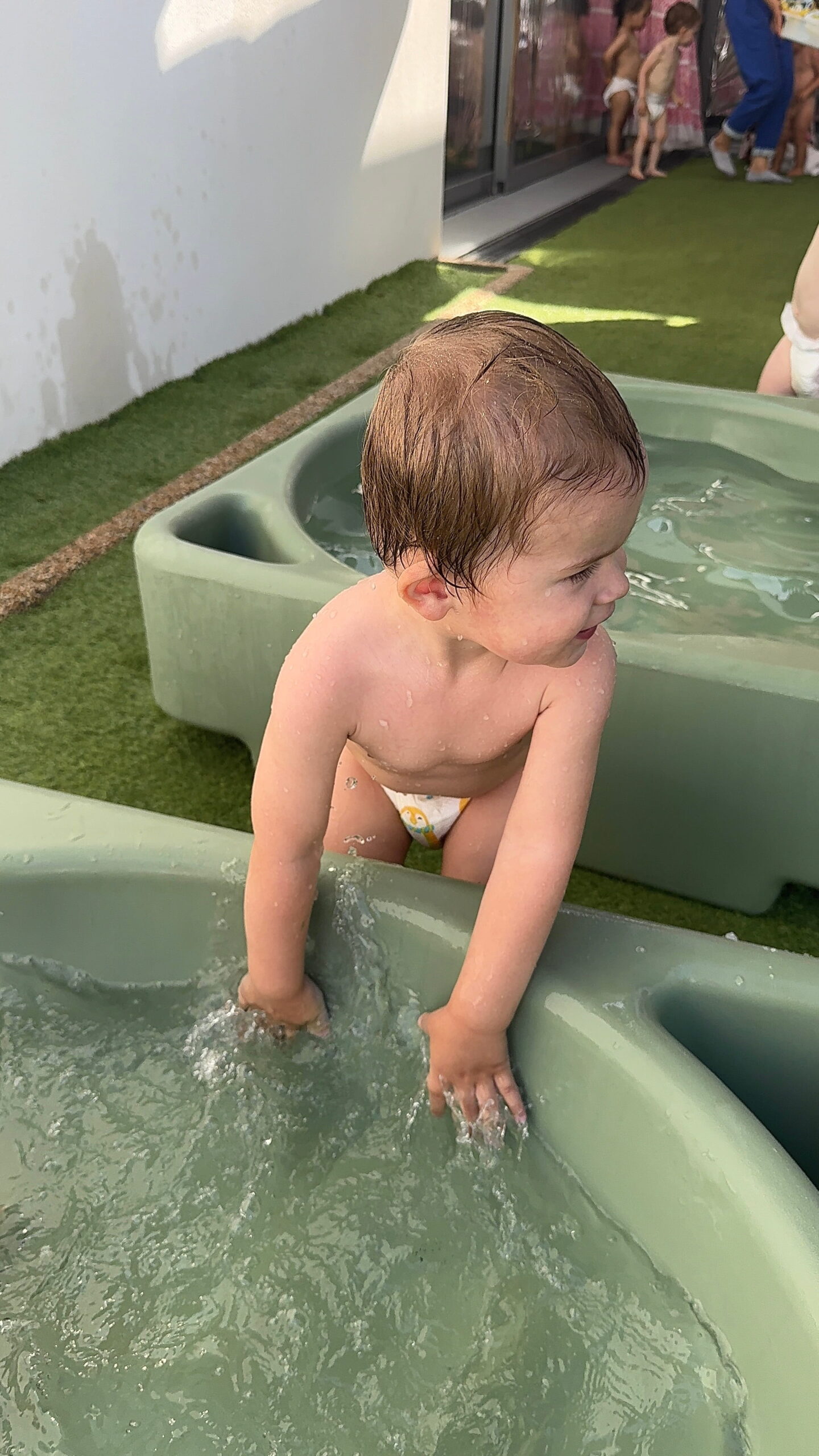 Un jeune enfant en couche splash ses mains dans un grand bassin d'eau vert sur une terrasse en gazon artificiel de la crèche Les Libellules de Vannes.