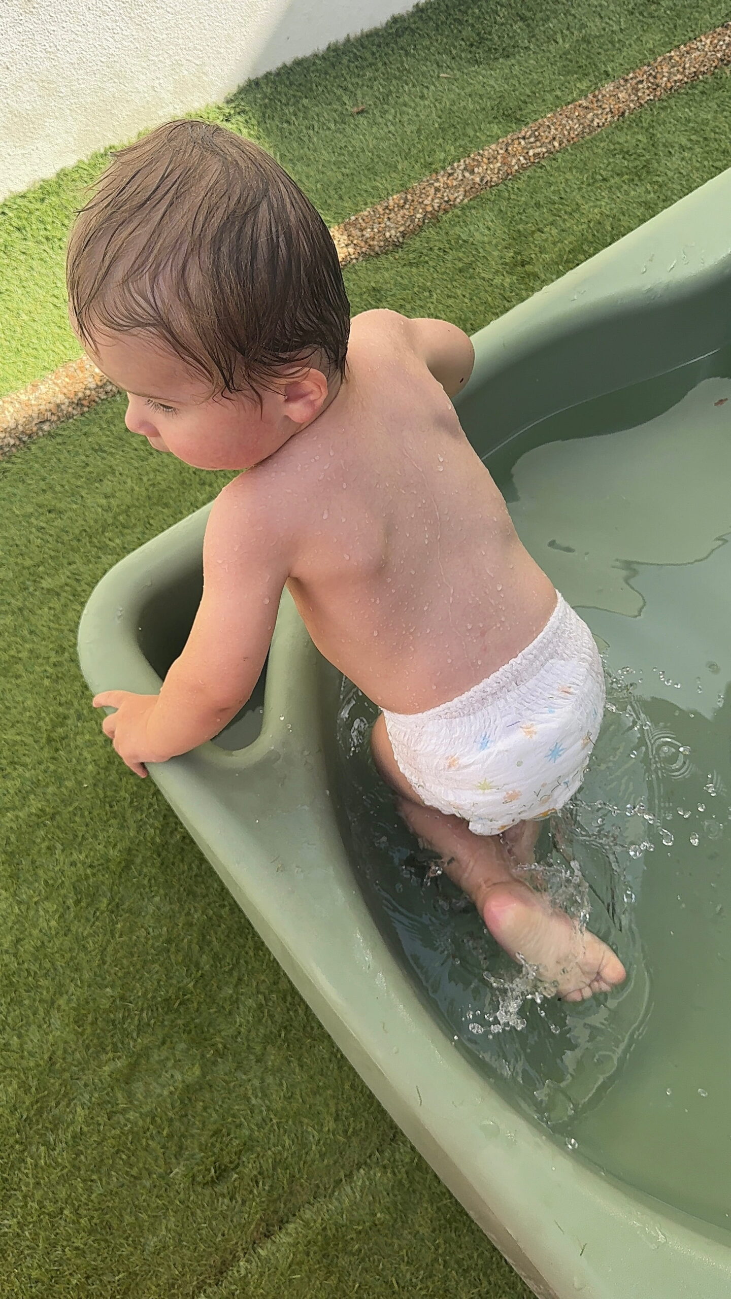 Vue de dos d'un jeune enfant en couche qui enjambe le bord d'une petite piscine en plastique vert clair sur la terrasse en gazon artificiel de la crèche Les Libellules à Vannes.