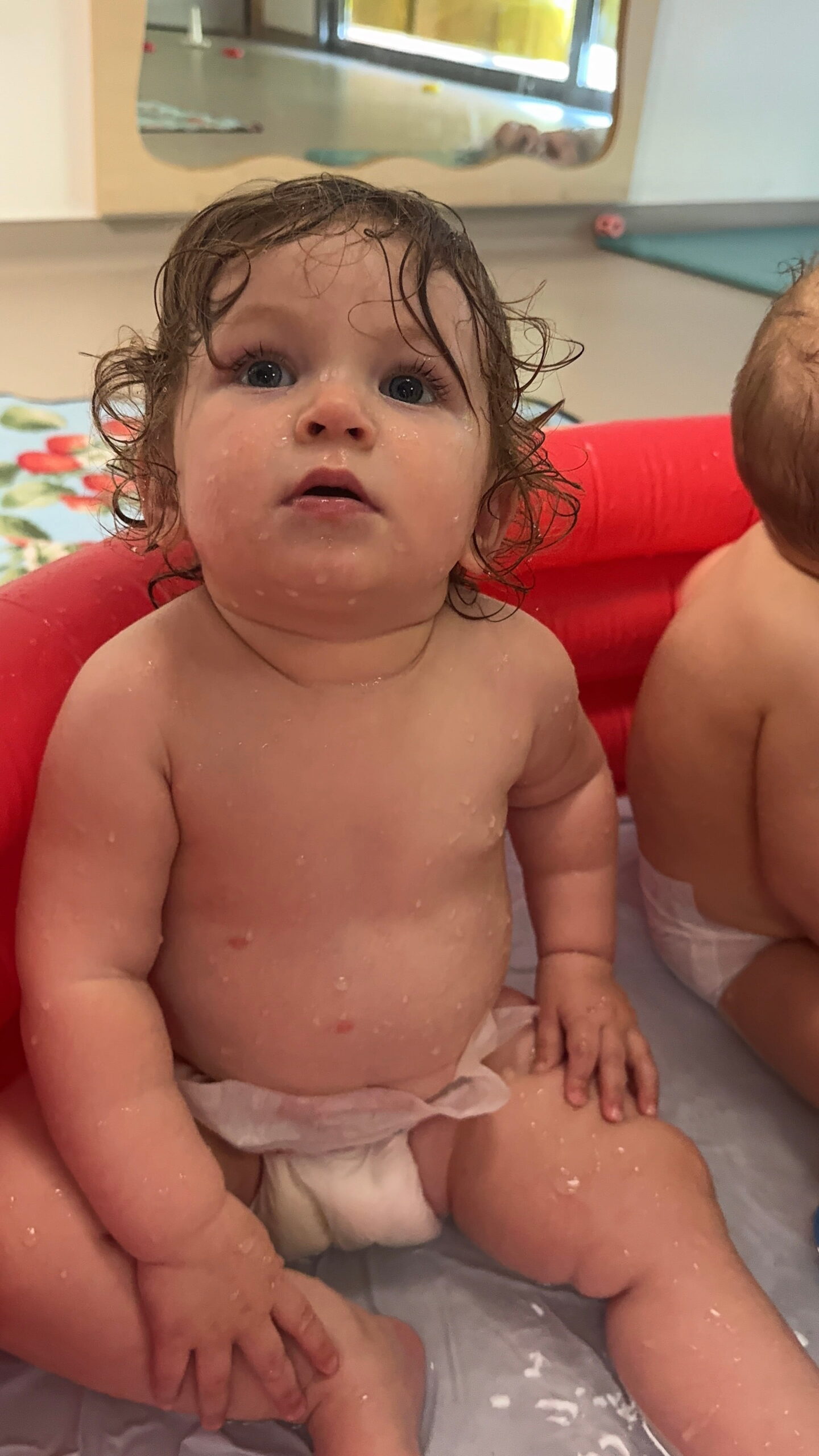 Un bébé aux cheveux bouclés et humides, portant une couche, est assis dans une petite piscine gonflable rouge à la crèche Les Papillons à Pontivy. Des gouttelettes d'eau perlent sur sa peau alors qu'il regarde vers le haut avec émerveillement. Un autre enfant est partiellement visible à ses côtés dans le bassin de la crèche Les Papillons.