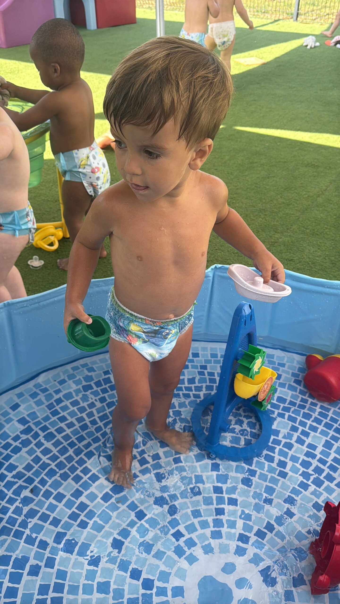 Un jeune enfant en couche de bain colorée se tient debout dans une piscine gonflable bleue à motif mosaïque à la crèche Les Libellules à Vannes. Il tient un gobelet vert dans une main et une petite baignoire jouet dans l'autre, entouré d'autres jouets d'eau et d'enfants en arrière-plan sur du gazon artificiel.