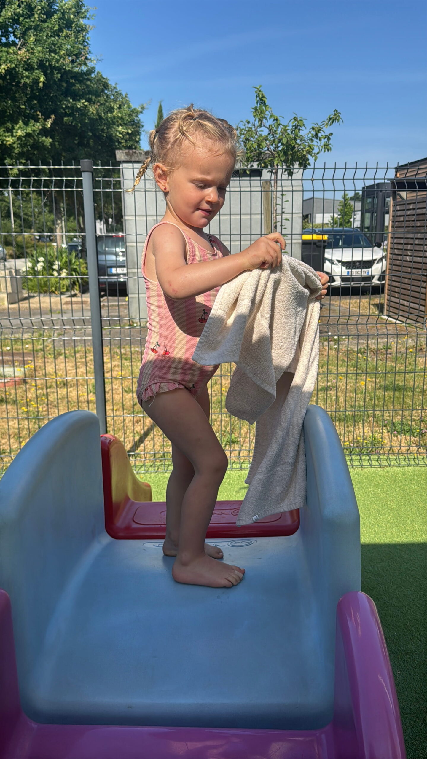 Une petite fille aux cheveux tressés, portant un maillot de bain rose à motif cerises, se tient debout sur un toboggan en plastique coloré à la crèche Les Libellules de Vannes. Elle tient une serviette beige et semble concentrée sur son pliage. L'arrière-plan montre l'extérieur de la crèche avec de la pelouse et une clôture.