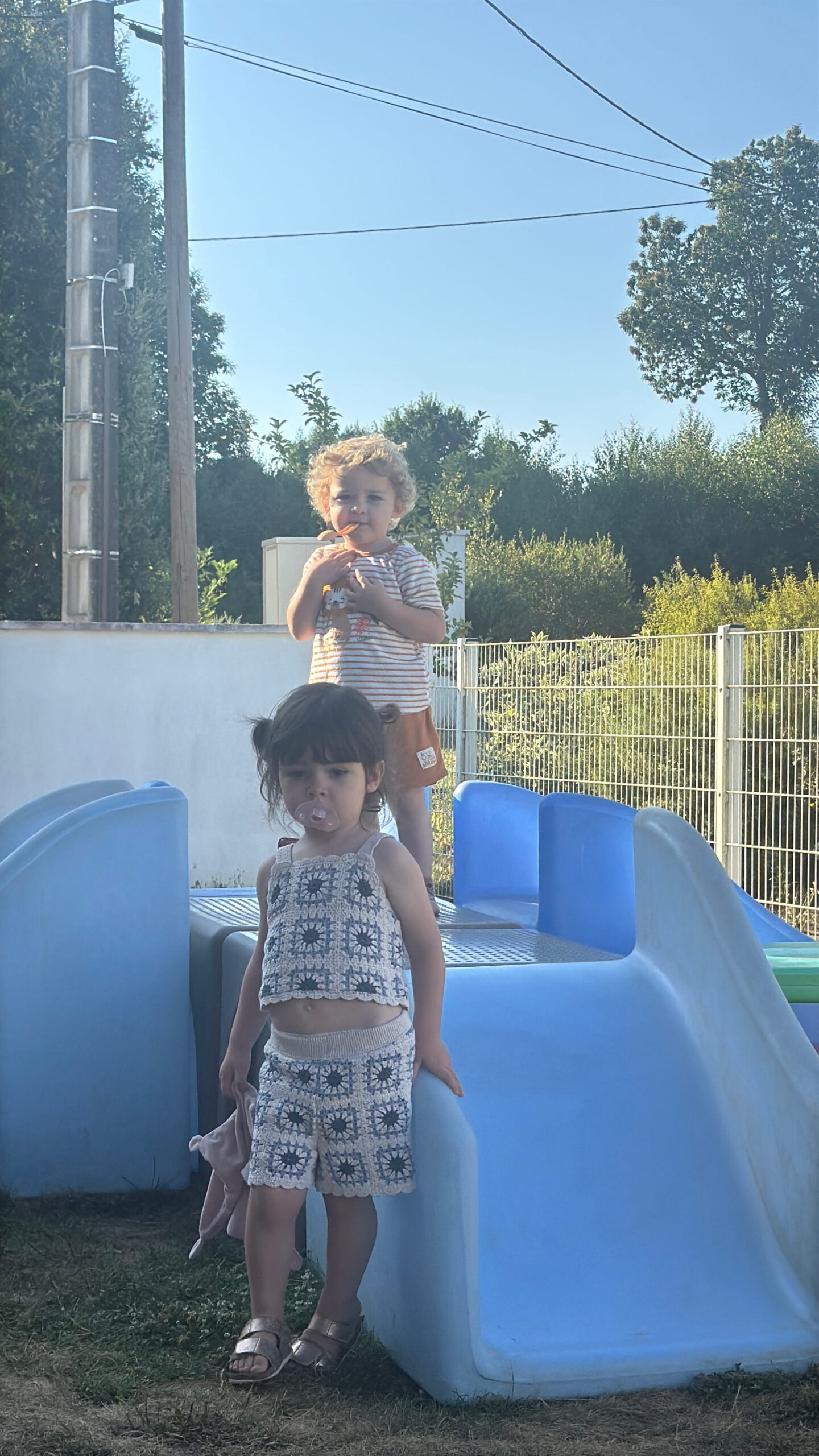 Une petite fille avec une tétine, vêtue d'un ensemble beige en crochet, se tient debout à côté d'un toboggan en plastique bleu dans le jardin de la crèche Les Papillons à Pontivy. Derrière elle, sur la structure de la crèche Les Papillons, un jeune garçon aux cheveux bouclés et en t-shirt rayé regarde l'objectif sous un grand soleil d'été.
