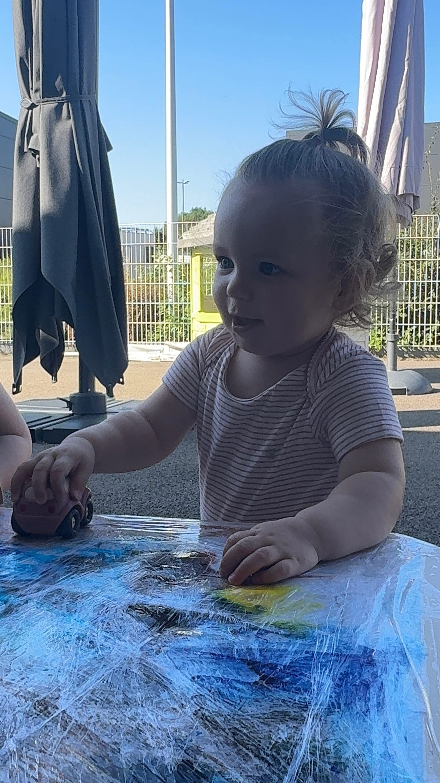 À la crèche Les Papillons à Pontivy, une petite fille avec un chignon haut sur la tête joue sur une table recouverte d'un film plastique protecteur et de peinture. Elle fait rouler une petite voiture en bois marron tout en souriant. Des parasols de la cour de la crèche Les Papillons sont visibles en arrière-plan sous un grand ciel bleu.