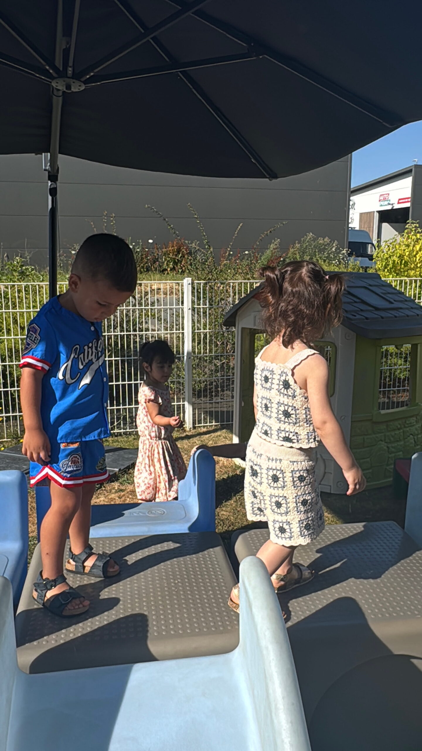 À la crèche Les Papillons à Pontivy, trois enfants s'amusent sur une structure de jeu extérieure abritée par un grand parasol noir. Un garçon en ensemble de baseball bleu regarde ses pieds, une petite fille en ensemble beige à motifs géométriques marche à ses côtés, et une autre fillette en robe fleurie se tient en arrière-plan près d'une cabane en plastique.