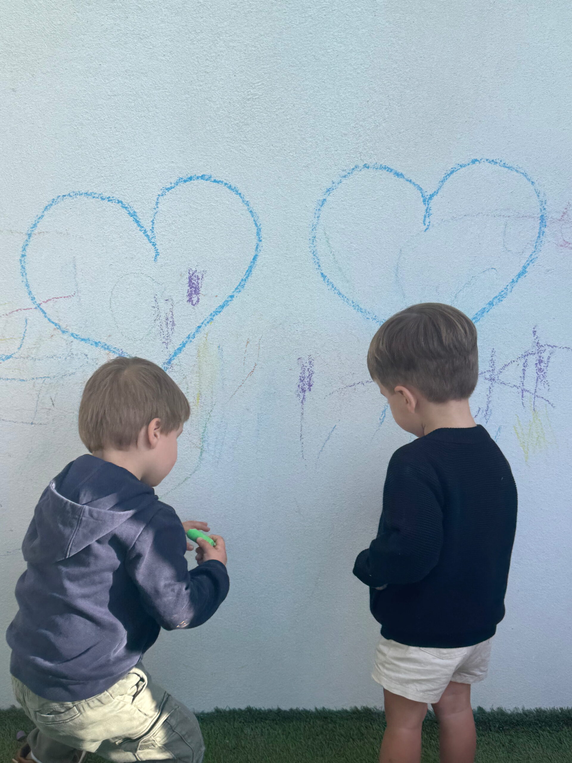 Deux jeunes enfants, vus de dos, dessinent à la craie sur un mur extérieur bleu clair. Le mur est orné de deux grands cœurs dessinés en bleu. L'un des enfants tient une craie verte.